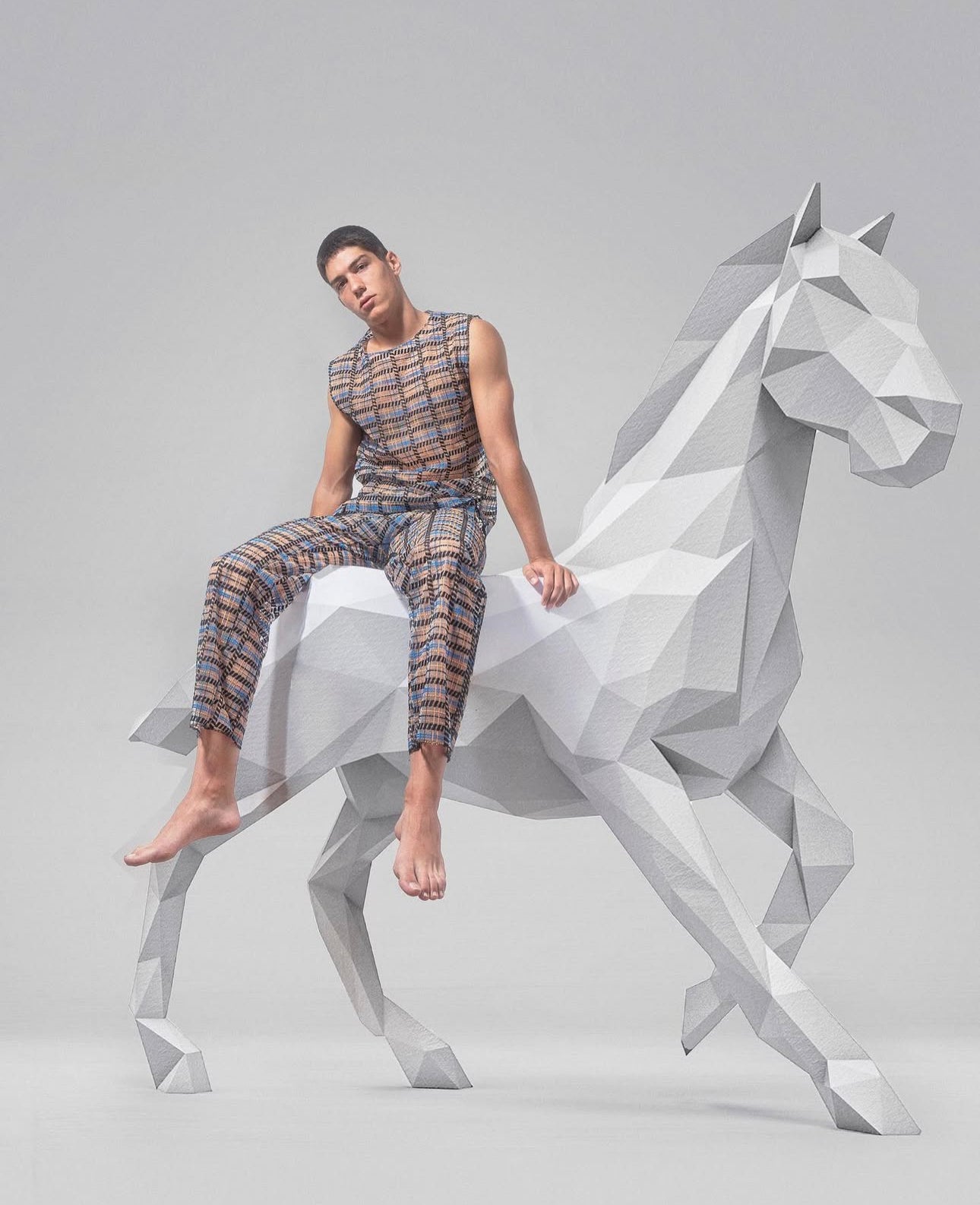 Barefoot man in earth-tone checkered outfit seated on a large white geometric horse sculpture, set against a minimal light gray background.