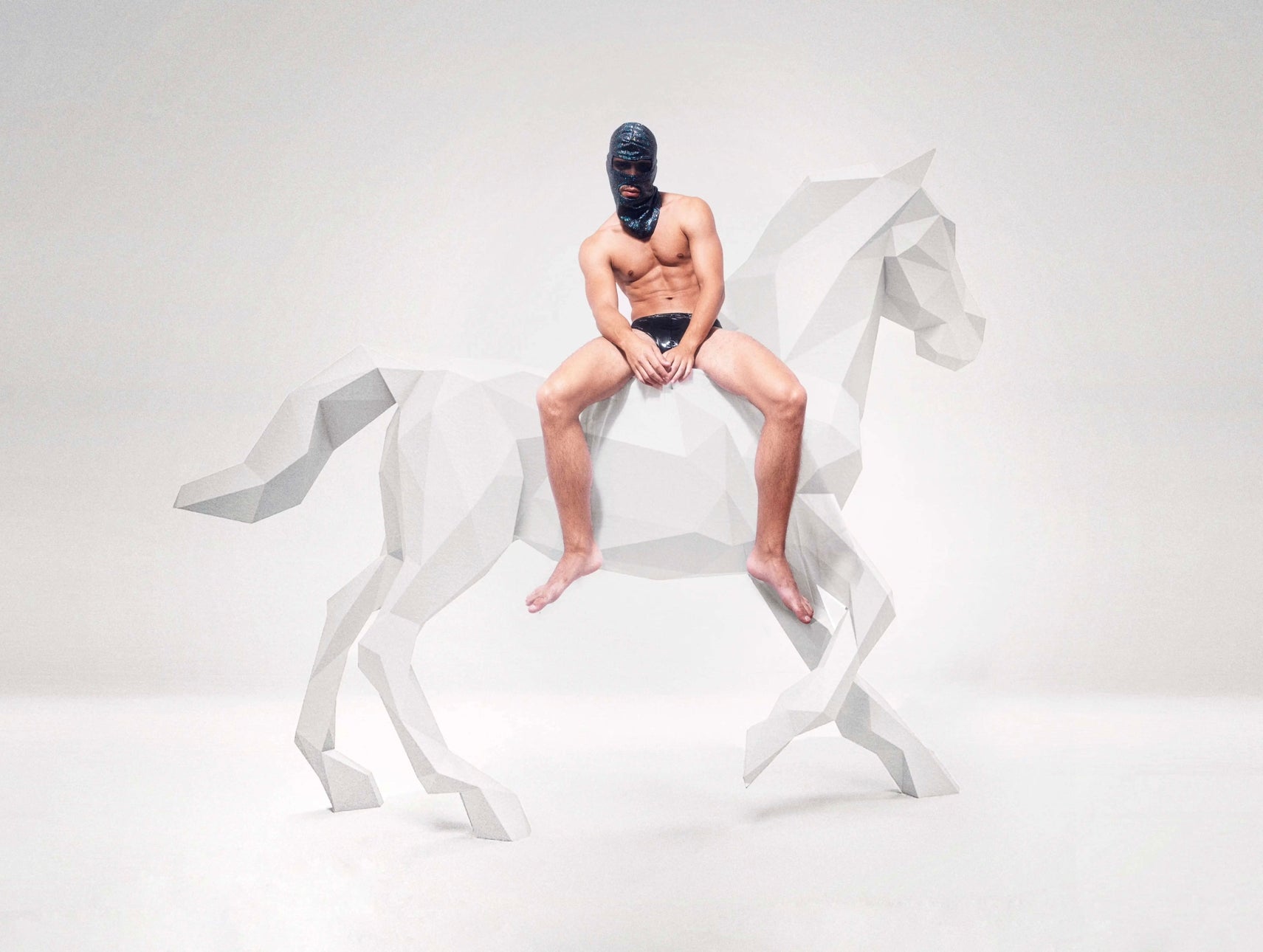 A man wearing a black balaclava and black underwear is sitting on a geometric, low-poly style sculpture of a horse. The horse is white and appears to be made of angular, faceted shapes. The background is a plain, light color, which emphasizes the contrast between the man and the horse sculpture.