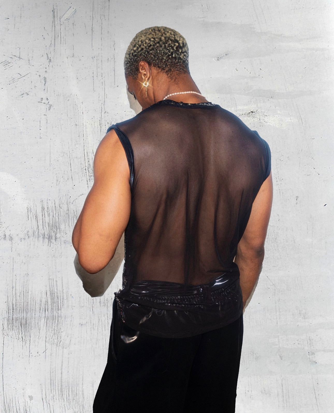 A person with short, curly hair dyed blonde is facing away from the camera. They are wearing a sleeveless, sheer black top that reveals their back. The top has a shiny, leather-like trim around the edges. The person is also wearing a pearl necklace and a hoop earring in their left ear. The background is a textured, light-colored wall.