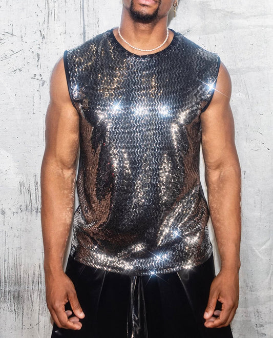 A man is wearing a sleeveless, shiny, sequined top that reflects light, creating a sparkling effect. He has a short beard and is wearing a small hoop earring in his left ear. Around his neck, there is a thin, beaded necklace. The background is a textured, light-colored wall with some abstract markings. His arms are muscular and he is standing with his hands relaxed by his sides.
